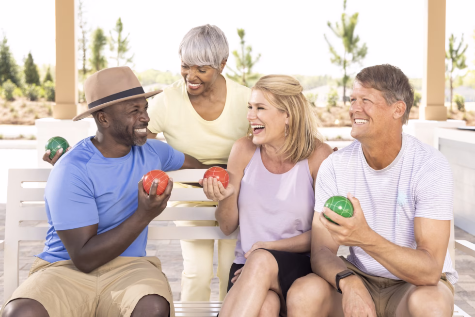 Four people enjoying an outdoor game, sitting on a bench, smiling and holding colorful balls, surrounded by trees on a sunny day.