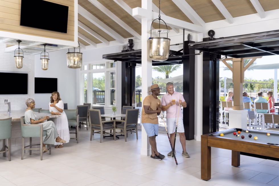 Four people socialize in a bright communal space with a pool table, bar seating, and large windows. Relaxed, inviting atmosphere indoors and outdoors.