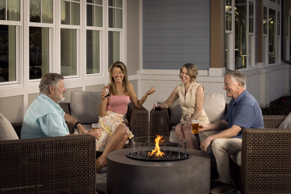 Four people enjoy drinks around a cozy outdoor fire pit, seated on wicker furniture by a house with large windows.