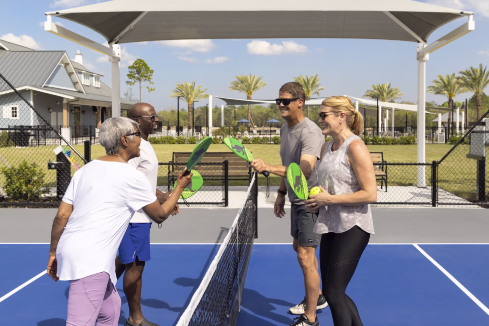 Four people enjoy a pickleball game on a sunny day, exchanging smiles and high-fives at the net. Palm trees in the background.