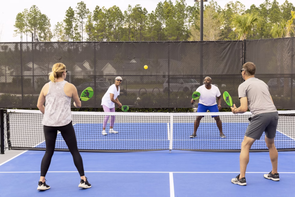 Four people playing pickleball on an outdoor court, surrounded by a mesh fence and trees, under a clear sky.