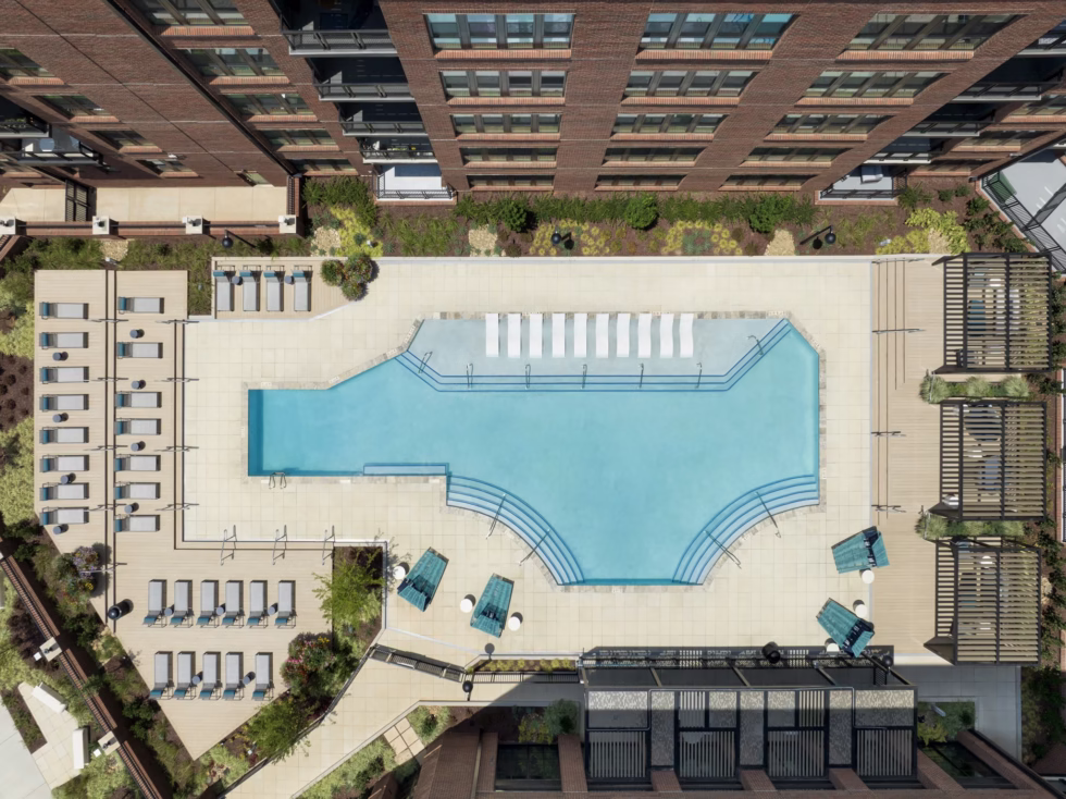 Aerial view of a modern apartment complex's outdoor pool surrounded by lounge chairs and greenery, with adjacent buildings visible.