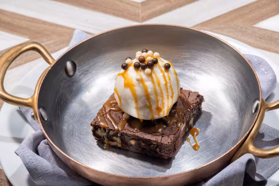 A brownie topped with ice cream, caramel drizzle, and chocolate pearls served in a metal pan on a plate with a cloth napkin.