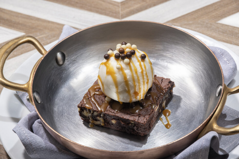 A brownie topped with ice cream, caramel drizzle, and chocolate pearls served in a metal pan on a plate with a cloth napkin.