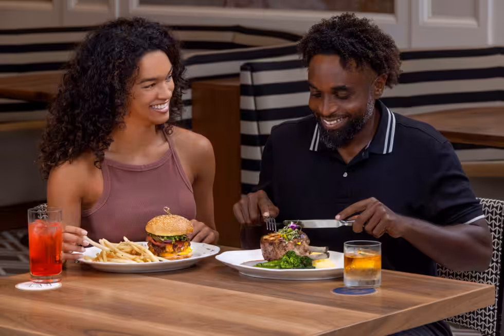 Two people dining in a restaurant, smiling while enjoying a meal of steak, burger, and drinks at a wooden table.