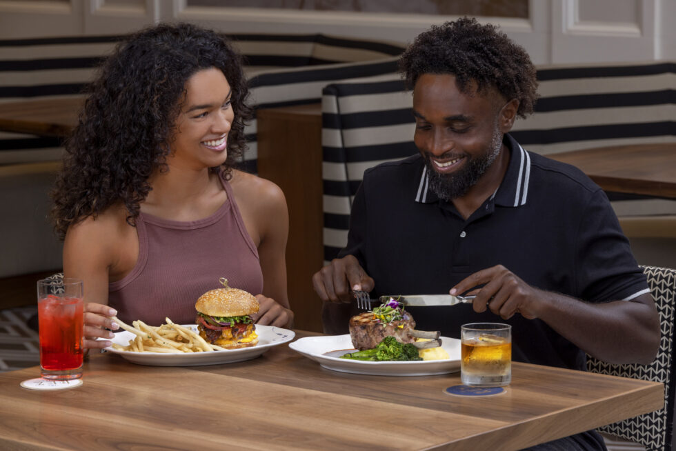 Two people dining in a restaurant, smiling while enjoying a meal of steak, burger, and drinks at a wooden table.