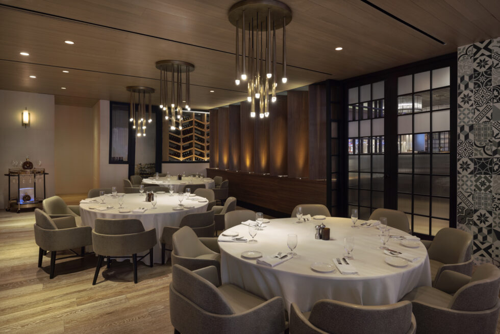 Elegant restaurant interior with round tables, modern chandelier lighting, and a wine rack; wooden and patterned elements accentuated by soft, ambient lighting.
