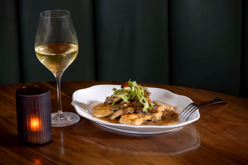 A table setting with a glass of white wine, candle, and a plated dish garnished with greens on a wooden surface.