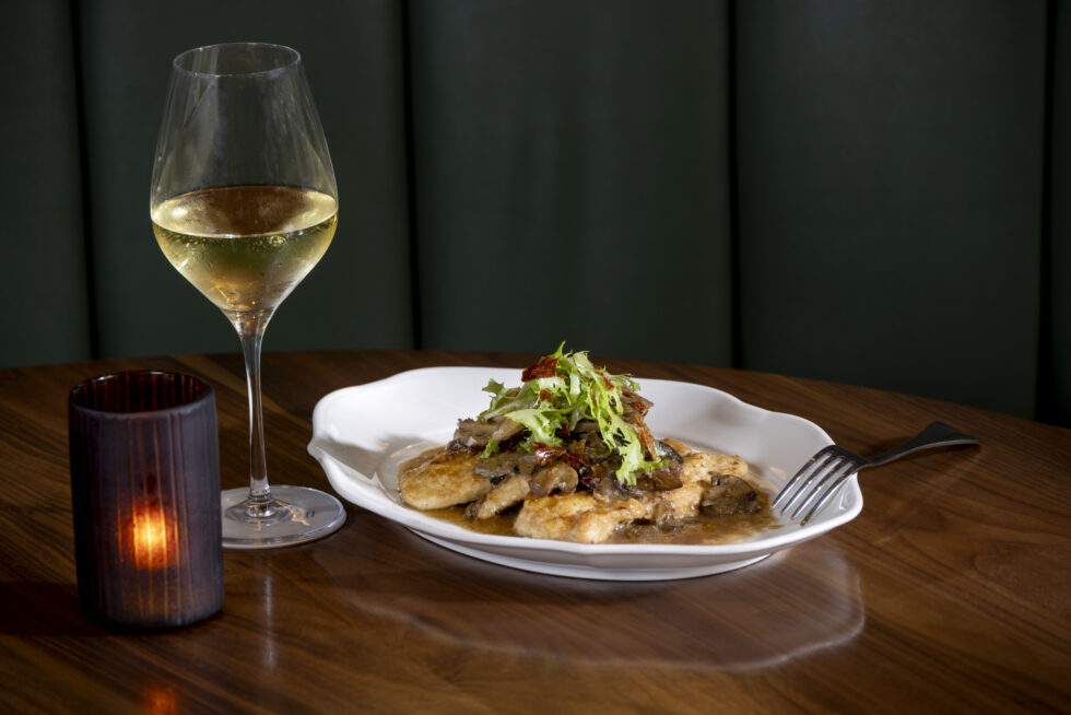 A table setting with a glass of white wine, candle, and a plated dish garnished with greens on a wooden surface.