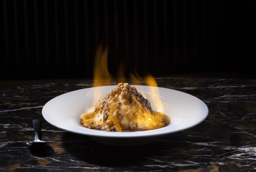 A flaming dessert is served on a marble table with a spoon placed nearby. The dish emits bright, lively flames.