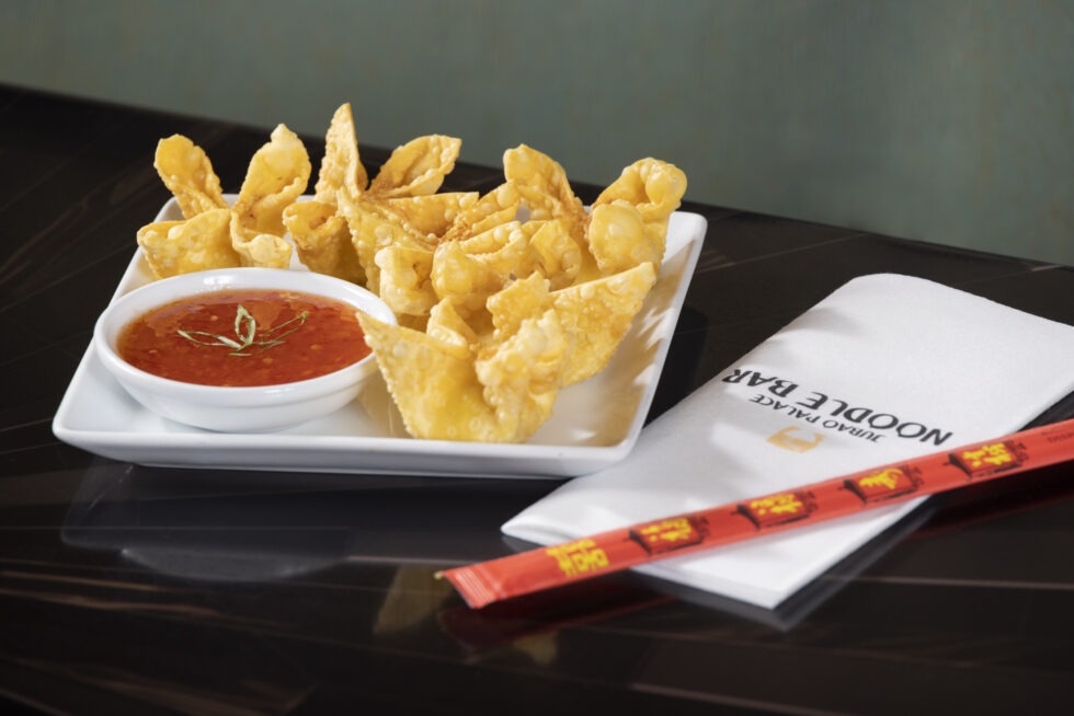 A plate of fried wontons with dipping sauce, chopsticks, and a napkin on a dark table.