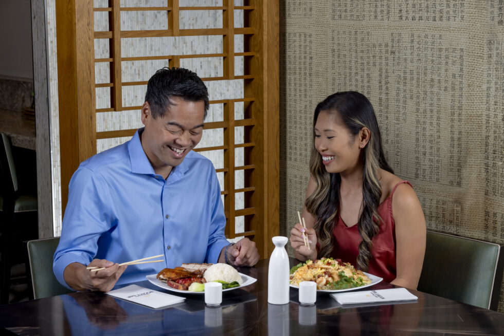 Two people enjoying a meal with chopsticks in a restaurant setting, surrounded by decorative wooden panels and patterned walls.