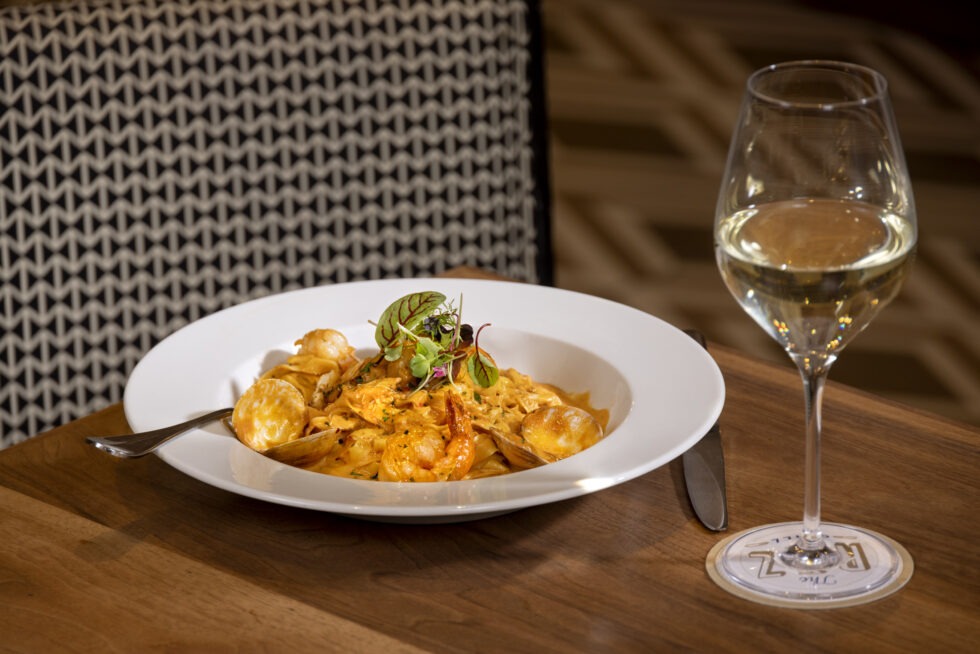 A plate of shrimp pasta garnished with greens beside a glass of white wine on a wooden table in a restaurant setting.
