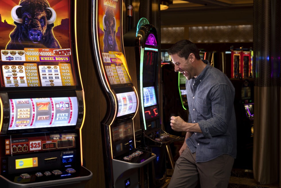 A person celebrates while playing a slot machine inside a casino. The machine displays winning combinations and jackpot amounts.