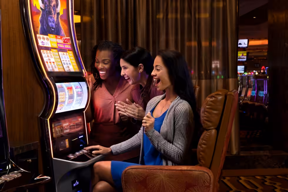 Three people excitedly play at a slot machine in a lively casino environment, surrounded by other gaming machines and dim lighting.