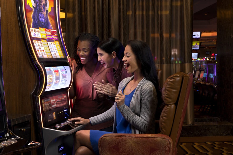 Three people excitedly play at a slot machine in a lively casino environment, surrounded by other gaming machines and dim lighting.