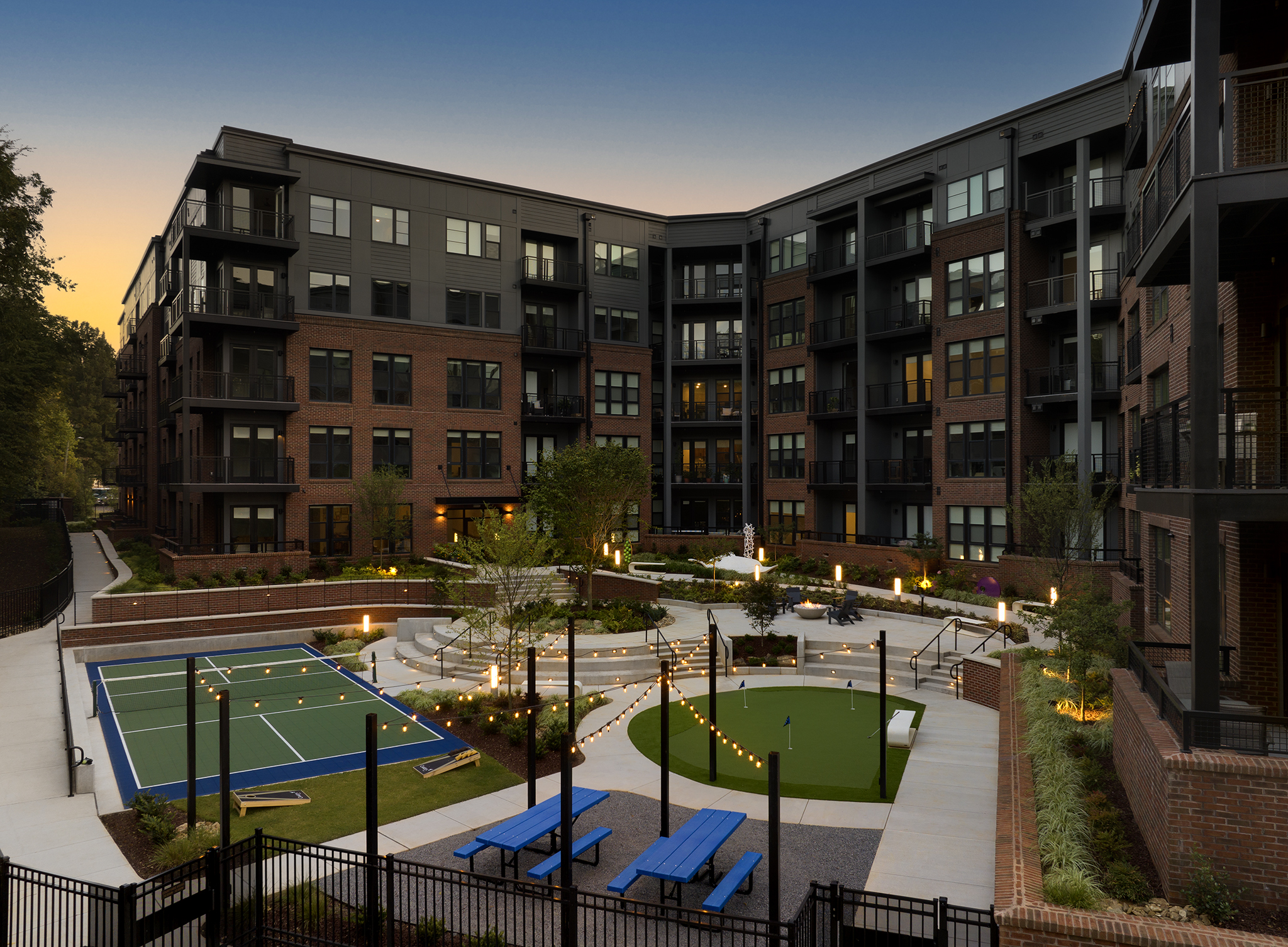A modern apartment complex courtyard features a sports court, seating areas, string lights, and green landscaping illuminated at sunset.